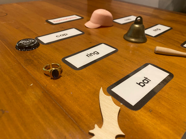 A wooden tabletop with several labeled cards and small objects arranged on it, to illustrate homonyms including a pink cap, a brass bell, a gold ring, a black bottle cap, and a light wood bat. The objects are photographed close up to show detail and interest.
