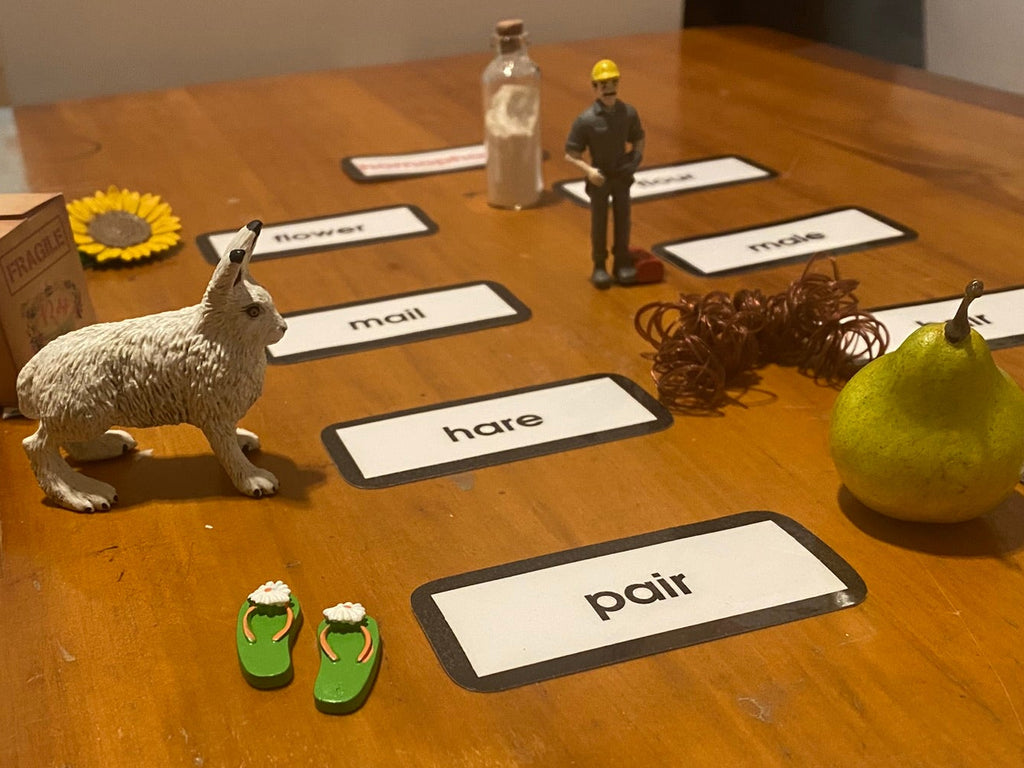 A wooden table set up to display a language-learning homophone activity with labeled cards, small figurines, a hare, a sunflower, a pear, and other props to be used to introduce the Homophone Key Experience.