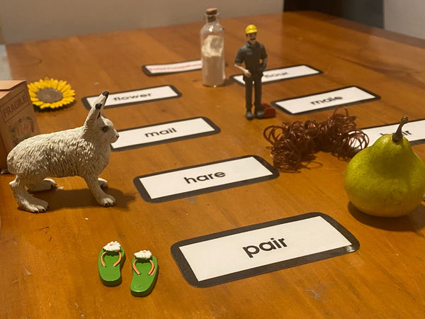 A wooden table set up to display a language-learning homophone activity with labeled cards, small figurines, a hare, a sunflower, a pear, and other props to be used to introduce the Homophone Key Experience.