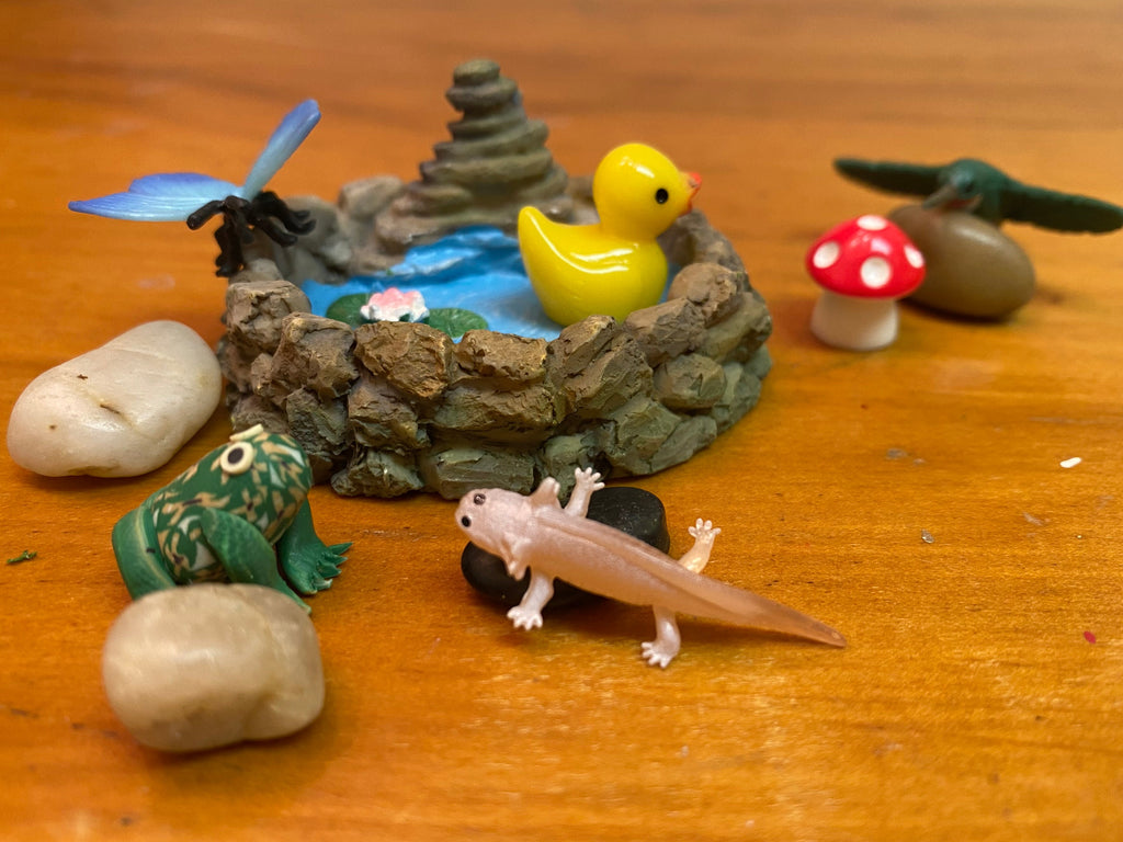 Small figurines of a duck, frog, butterfly, hummingbird, and axolotl on a wooden surface with decorative rocks, mushroom and pond background.