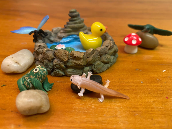 Small figurines of a duck, frog, butterfly, hummingbird, and axolotl on a wooden surface with decorative rocks, mushroom and pond background.