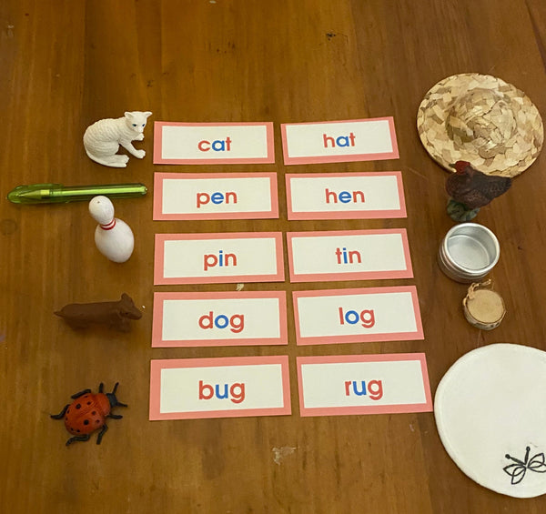 A wooden tabletop with a set of educational word cards arranged in rows, alongside small toy objects in rhyming pairs.